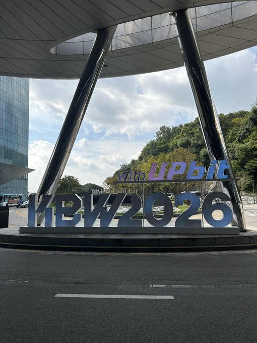 A sign reading 'KBW2026 with Upbit' is displayed beneath a modern architectural canopy structure.