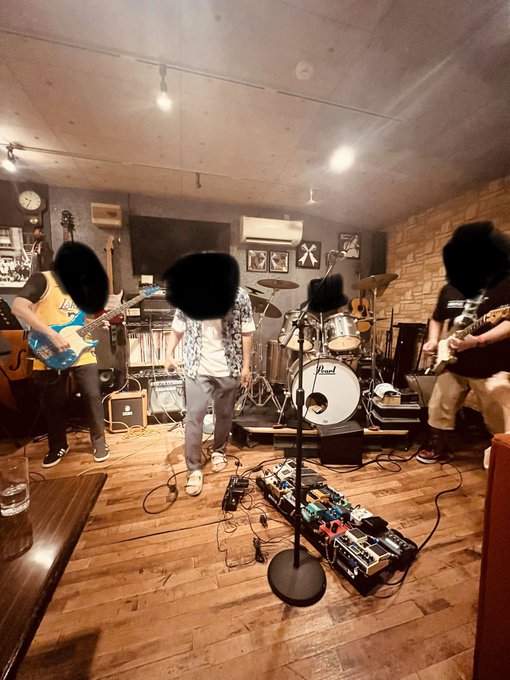 A band performs live in an intimate wooden-floored venue, with members playing guitar and drums around a Pearl drum kit and an array of effect pedals, while a microphone stand sits center stage. The faces of the performers have been blacked out for privacy, matching the poster's playful note that faces can't be shown since there are other members besides themselves.