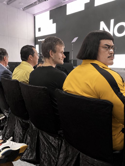 Four people seated in a row wearing black velvet chairs, with the person on the right in a yellow and black outfit and a distinctive bowl haircut.