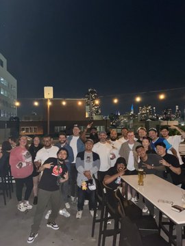 A nighttime group photo of people gathered on a rooftop patio with string lights and a city skyline in the background.