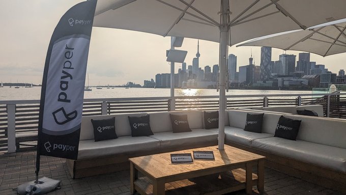 An outdoor rooftop lounge area with white sofas, branded 'payper' pillows, and a tall 'payper' banner, overlooking a city skyline with a prominent tower across the water.