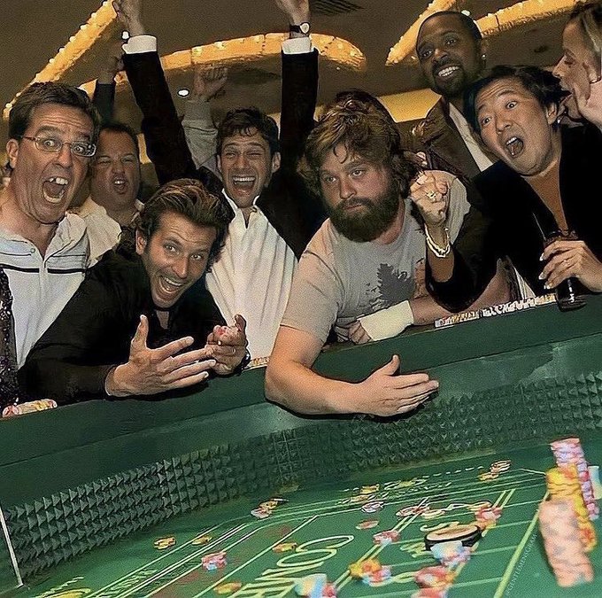 A group of men react with wild excitement—arms raised, mouths open in cheers—around a green craps table stacked with colorful chips, capturing the euphoric energy of a big gambling win that matches the post's boast about "turning the $10M into $20M."