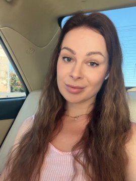 A selfie of a woman with long brown hair wearing a pink and white striped top, sitting inside a car with a window and greenery visible in the background.