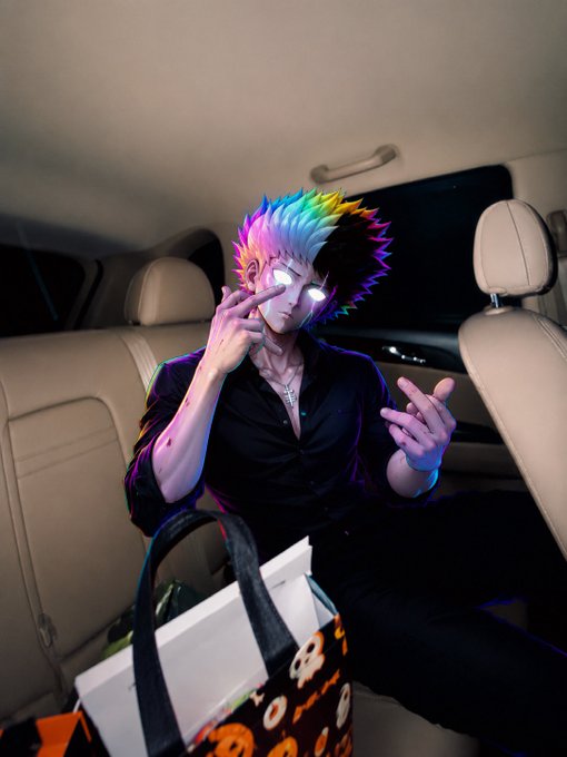 An anime-style character with rainbow hair and glowing white eyes sits in the back of a car at night, wearing a black shirt and crossing their fingers while a Halloween-themed bag rests on their lap.