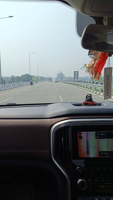 A view from inside a car driving on a highway, showing a road stretching ahead with streetlights, a small religious figurine and decorative tassels on the dashboard, and an 'INVOLVE' air freshener hanging from the rearview mirror.