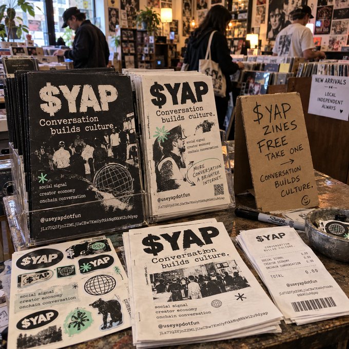 A retail display of '$YAP' branded zines, stickers, and merchandise with the tagline 'Conversation builds culture' sitting on a counter in what appears to be a record or book store.