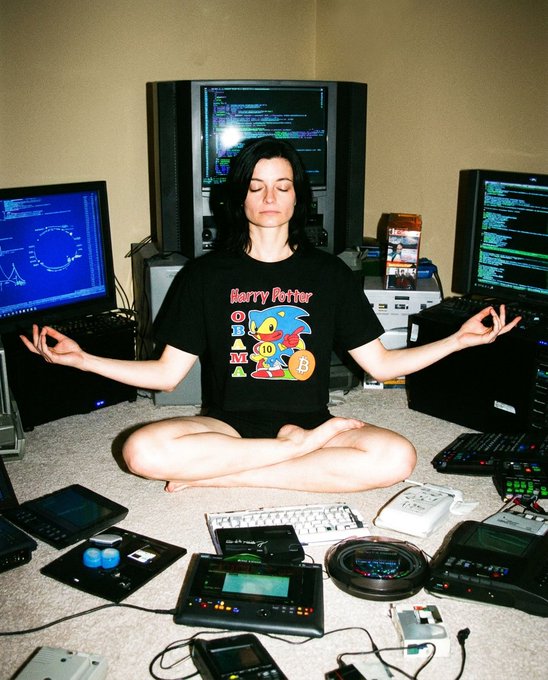 A woman sits cross-legged in a meditative pose with her eyes closed, surrounded by an array of vintage computer monitors, keyboards, and electronic devices, wearing a black t-shirt featuring 'Harry Potter' and a Sorting Hat with the houses H, B, M, and a Bitcoin logo.