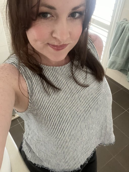 A selfie of a woman with brown hair wearing a sleeveless black-and-white striped top, standing in what appears to be a bathroom with a green towel visible in the background.