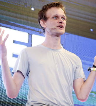 The image shows a man wearing glasses and standing next to a blue screen with a large white letter "E". He appears to be giving a presentation or speech, as his hands are raised in the air during this moment. The background of the image is blurred, but one can make out that it's likely an indoor setting. There is also some text present in the image that reads: "Just In: ETHEREUM FOUNDATION RE-STRUCTURES, CUTS 20% OF WORKFORCE The Ethereum Foundation is restructuring its core research arm, rebranding R&D to "Protocol" and narrowing focus to L1 scaling, blobs and UX.". This additional information provides context about the setting or subject matter being discussed by the man in the image.