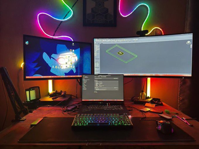 The image shows a desk setup with two computer monitors, one larger than the other, accompanied by a laptop. The screens display a cartoon character from "Frens", which appears to be a popular video game. A keyboard and mouse are placed in front of each monitor for comfortable use. There is also a cup on the desk, possibly containing coffee or tea. The overall scene suggests that someone might have been working late into the night or enjoying some leisure time playing video games with friends online.