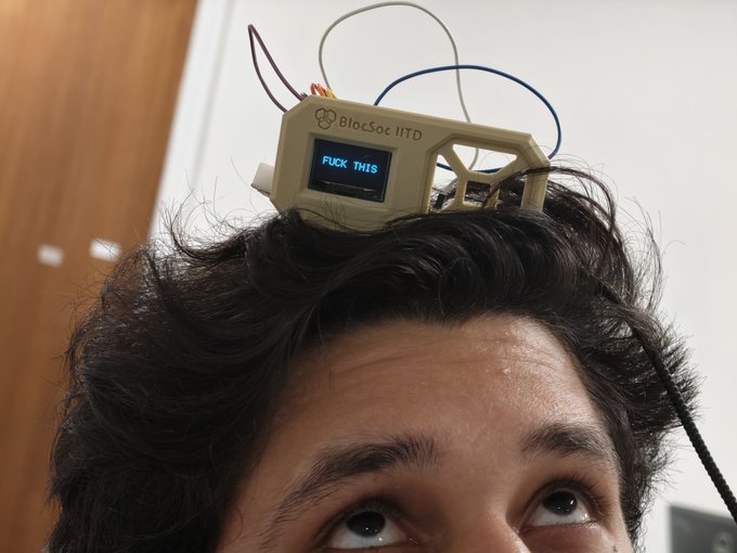 A close-up of a person's forehead with a small electronic device labeled 'BlocSoc IITD' mounted on their head, its screen displaying the text 'FUCK THIS'.