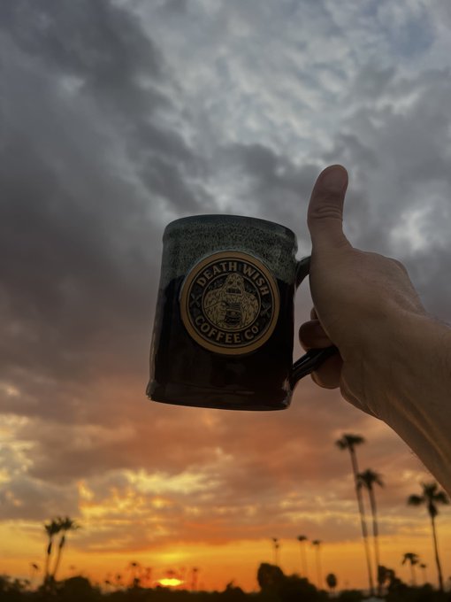 A hand holds a black ceramic mug bearing the 'DEATH WISH COFFEE CO.' logo against a backdrop of a cloudy sunset sky.