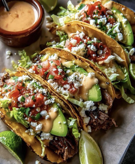 A row of crispy street-style tacos filled with tender steak, fresh avocado slices, pico de gallo, crumbled cotija cheese, and shredded lettuce, drizzled with a creamy chipotle sauce and accompanied by lime wedges. The vibrant presentation perfectly matches the post's "minted steak tacos" description, though the accompanying street corn and spicy elements aren't visible in the frame.