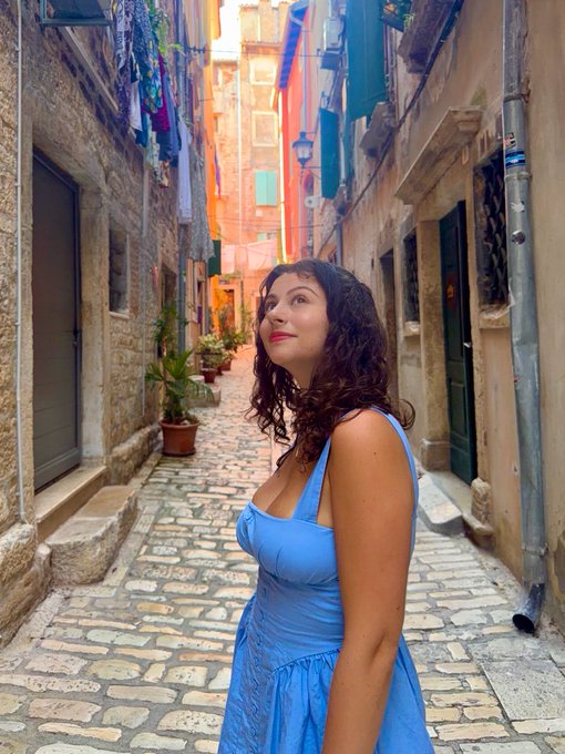 A young woman in a blue sundress poses in a narrow, sunlit cobblestone alleyway lined with colorful laundry overhead and weathered stone buildings. The image perfectly complements the post about choosing "the road less traveled," as she explores a charming, off-the-beaten-path Mediterranean side street far from typical tourist routes.