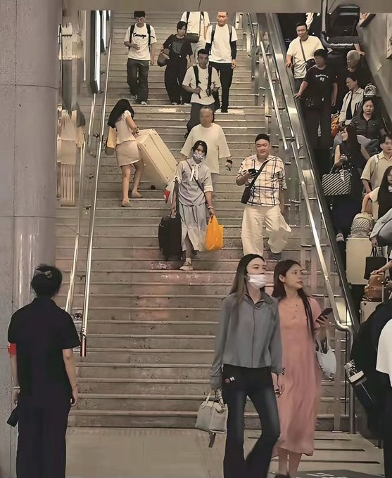 A busy crowd of people, some carrying luggage, descends a wide staircase and rides an escalator in what appears to be a transit station or airport.