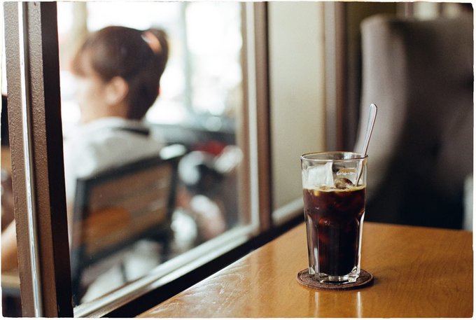 A glass of iced coffee with a spoon sits on a wooden table at a window-side seat, while a person with their hair up sits blurred in the background. The scene captures a quiet moment of reflection at a café, fitting the post's message of staying focused amid unpredictable markets.
