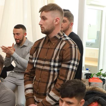 A group of men sit together, with one wearing a brown checkered Burberry shirt clapping his hands.