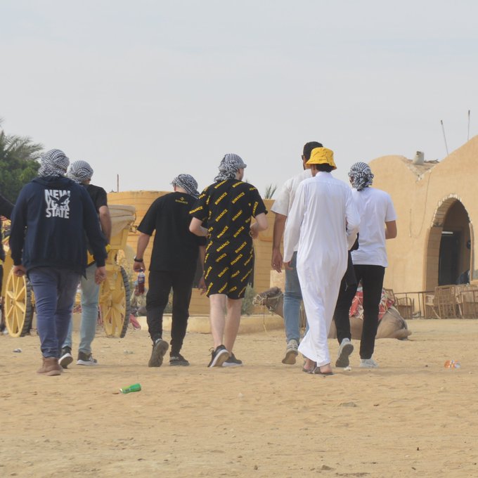 Group of people wearing keffiyehs and casual clothing walking together across a sandy desert area with traditional mud-brick structures in the background.
