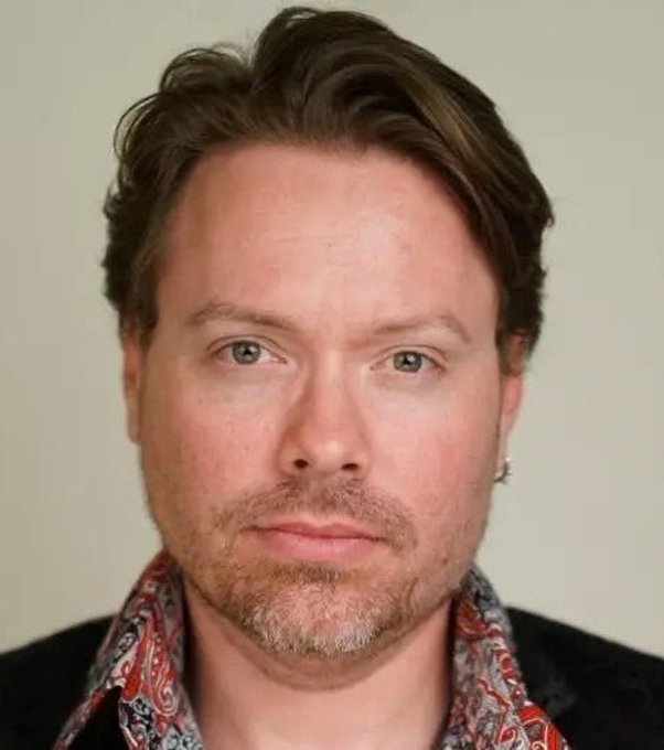 A close-up portrait of a man with brown hair, a light beard, and blue eyes, wearing a patterned shirt under a dark jacket against a plain background.