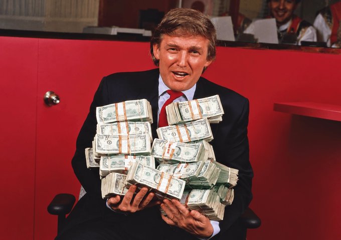 A man resembling Donald Trump, wearing a dark suit and red tie, holds a massive pile of banded US dollar bills while seated in front of a red cabinet.