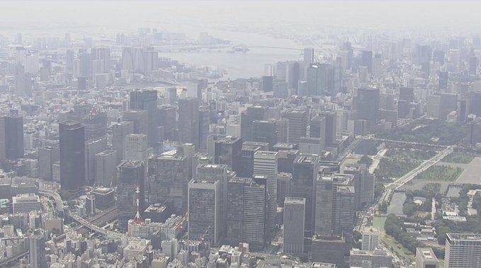 An aerial view of a hazy Tokyo skyline, featuring numerous skyscrapers and a large green park, likely represents the traditional financial infrastructure that JPYC is unaffected by disruptions from.