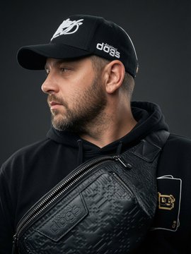 A man with a short beard wears a black cap with a "dogs" logo and black hoodie, with a textured black crossbody bag featuring a gold "DJ" buckle.