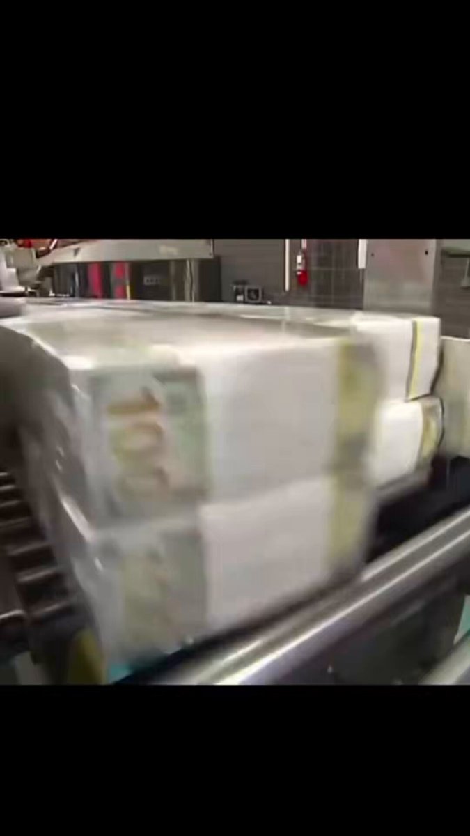 The image shows a stack of 100 dollar bills being processed by an automated machine, likely at a bank or financial institution. The money appears to be in pristine condition, indicating that they are new and unused. The scene takes place against a gray background, which contrasts with the vibrant colors of the money.