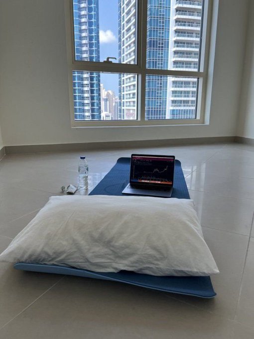 A laptop displaying a crypto trading chart sits on a bare blue mattress on the tiled floor of an empty high-rise apartment, with a water bottle nearby. The sparse, minimalist setup humorously illustrates the stereotype of a crypto trader living frugally while watching their investments.