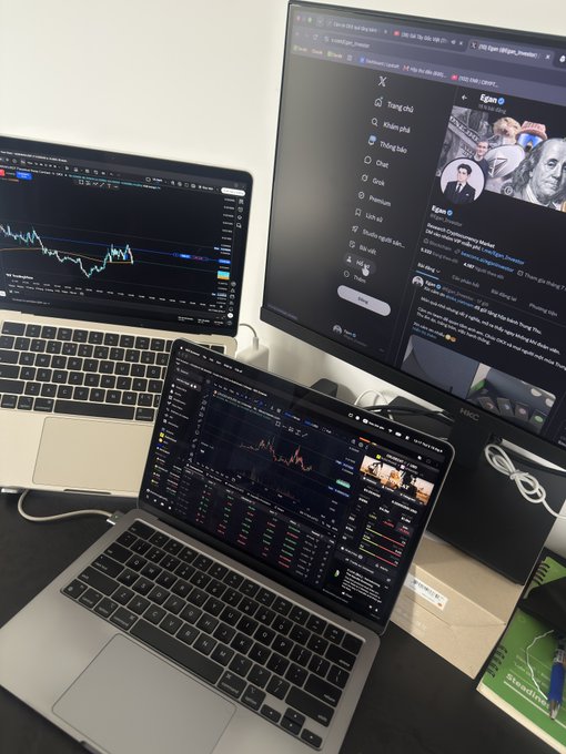 A multi-screen trading workstation displaying candlestick charts on a laptop and external monitor, alongside the X (Twitter) feed open to user 'Egan.' showing a post about Chevron.