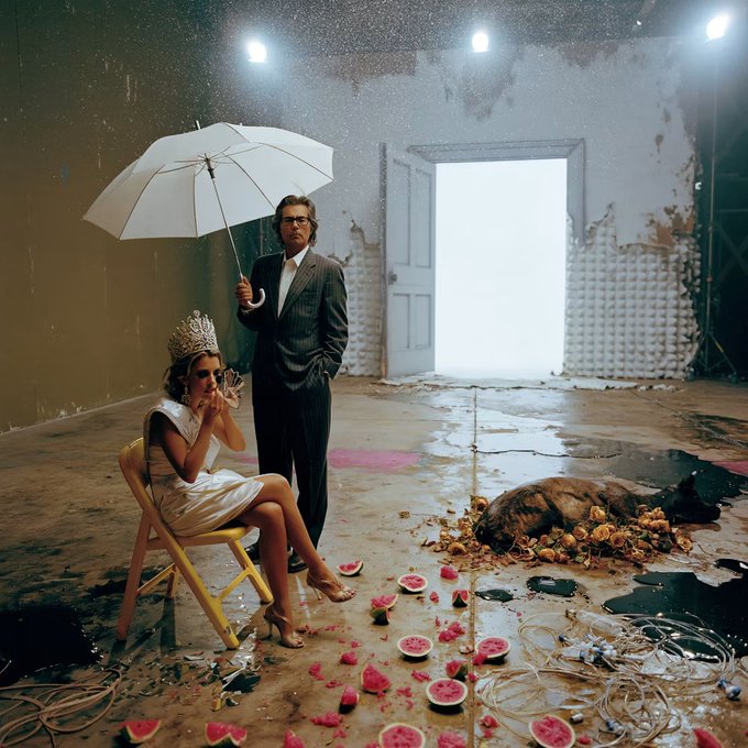 An artistic, moody photo shows a seated woman wearing a crown and holding a phone, while a man in a suit stands behind her holding a white umbrella, in a room with watermelons scattered on the floor.