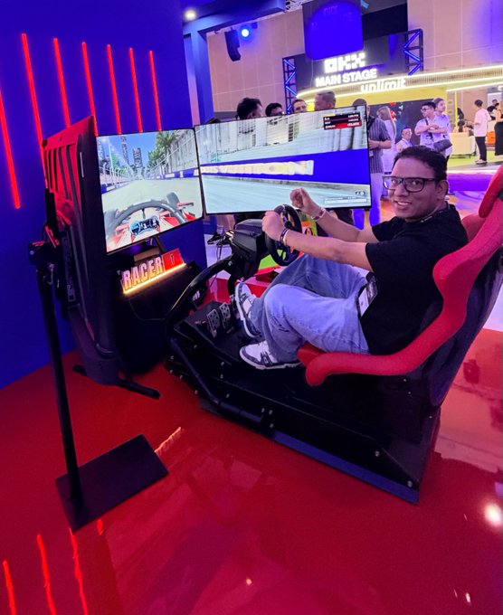A man wearing glasses and a black shirt sits in a "RACER 1" arcade racing simulator at the "MAIN STAGE #TOKEN2049," holding a steering wheel in front of dual monitors displaying a racing game. 

The image shows a lighthearted moment of relaxation at TOKEN2049 Singapore, contrasting with the post's warning to stay vigilant against social engineering attempts disguised as friendly networking opportunities—even casual encounters in spaces like this event can be exploited by scammers gathering personal information.