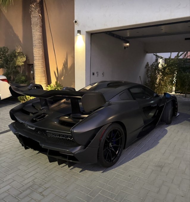 A matte black McLaren sports car with a large rear wing parked in the driveway of a modern home at dusk.