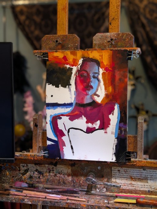 An artist's easel displaying a colorful, expressive portrait painting of a figure in red and blue tones, surrounded by paintbrushes and art supplies in a studio environment.
