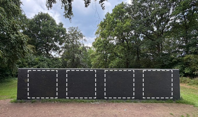 A long, black wall or structure set within a park-like area with green grass and large trees. The black surface features five rectangular frames outlined by white dashed lines.