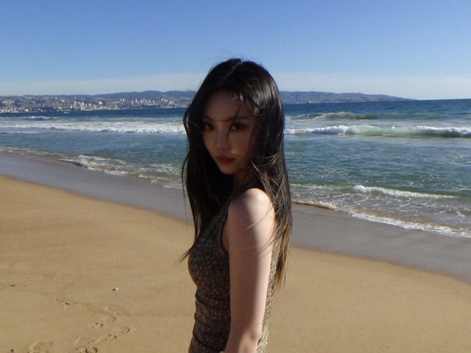 A woman with long dark hair stands on a sandy beach, looking back over her shoulder with the ocean and distant hills visible behind her.