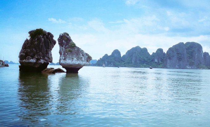 A scenic view of limestone karst rock formations rising from calm blue-green waters, likely Ha Long Bay in Vietnam, with small boats visible in the distance.