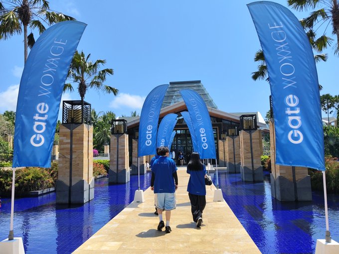 Two people in blue shirts walking down a pathway lined with blue 'Gate VOYAGE' flags toward a building entrance in a tropical setting.
