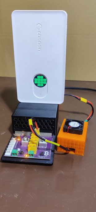 An image of a white 3D printer or electronic device with a green pixelated display, connected to an orange 3D-printed fan housing and other electronics.