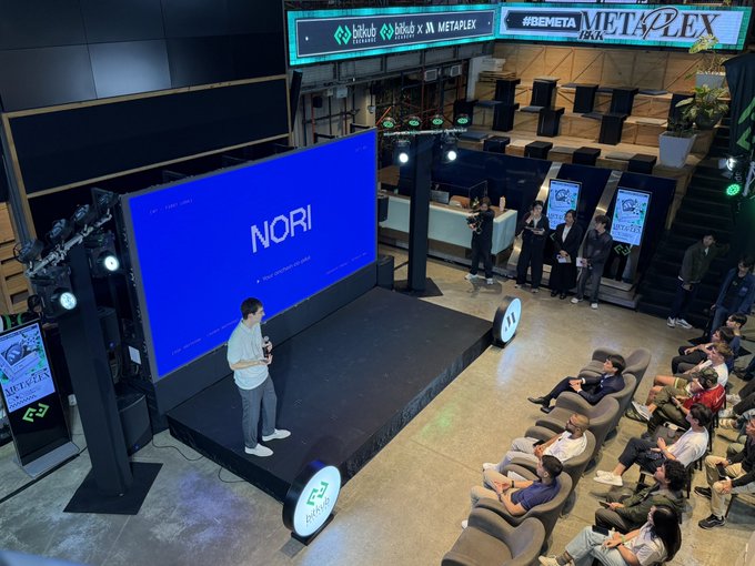A presenter on stage introduces "NORI" on a large blue screen during a MetaPlex event, with banners reading "#BEMETA" and "METAPLEX" visible above. The presentation aligns with the announcement of NORI as MetaPlex's official AI copilot.