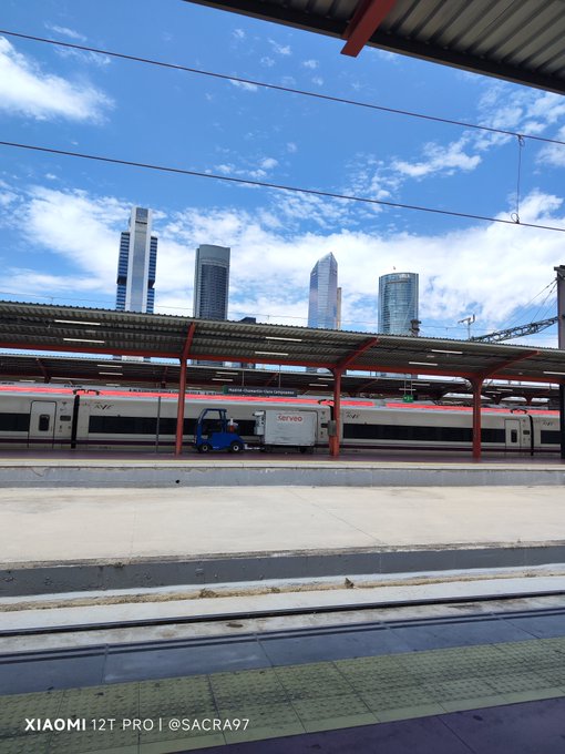 A photo taken from a train station platform showing a parked train with a 'Correos' (Spanish postal service) truck next to it, with tall skyscrapers in the background under a partly cloudy sky.