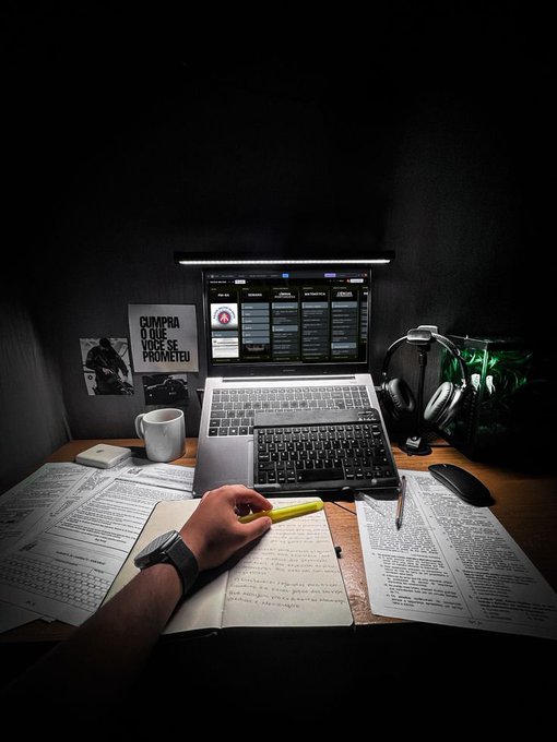 Moody photograph of a person studying at a desk with a laptop, external keyboard, headphones, scattered papers, and a Brazilian flag, with a poster reading 'CUMPRA O QUE VOCÊ SE PROMETEU' (Keep what you promised yourself).