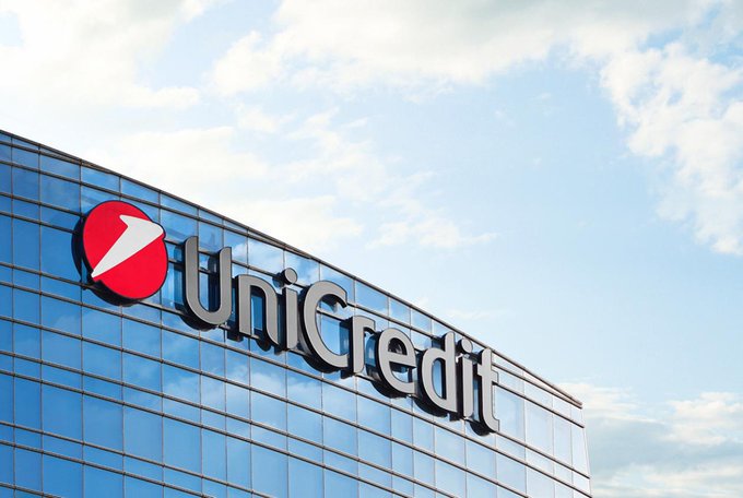 The UniCredit logo, featuring a red circle with a white sail design, mounted on the glass facade of a modern building against a partly cloudy sky.