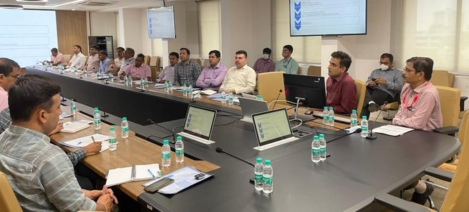 A group of people sit around a large conference table in a meeting room, with laptops, water bottles, and a presentation screen visible in the background.