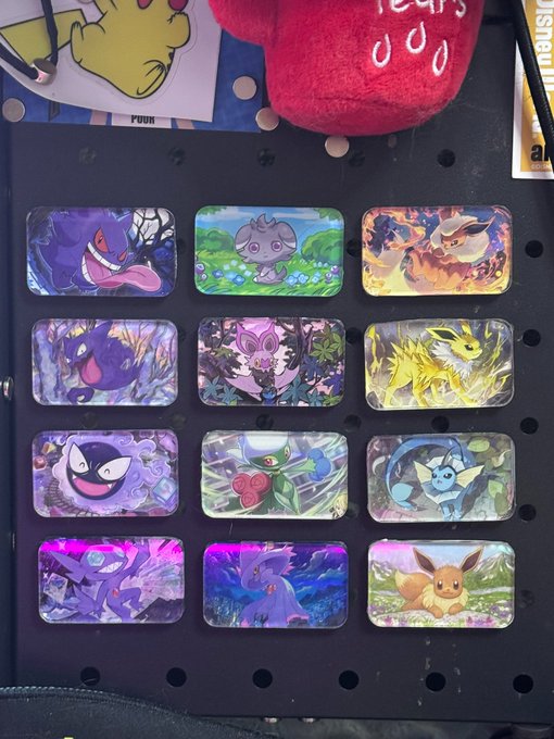 A collection of twelve holographic Pokémon-themed stickers featuring Gengar, Muk, Mimikyu, Gastly, Noivern, Jolteon, and Eevee, displayed in a pegboard case alongside a red plush toy, ready to inspire suggestions for the next design.