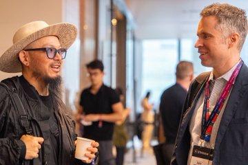 Two men in conversation at an event, one wearing a straw hat and glasses, the other in a suit with a red lanyard.