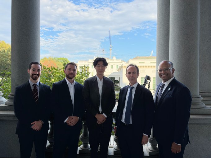 Five men in business suits pose together on a columned balcony of a stately building, with the context indicating Injective CEO Eric Chen and his delegation meeting with U.S. White House, SEC, and CFTC officials to discuss compliant onchain markets. The formal, official setting of the photo visually reinforces the article's theme of Injective's push into institutional finance and regulatory engagement around tokenized real-world assets.