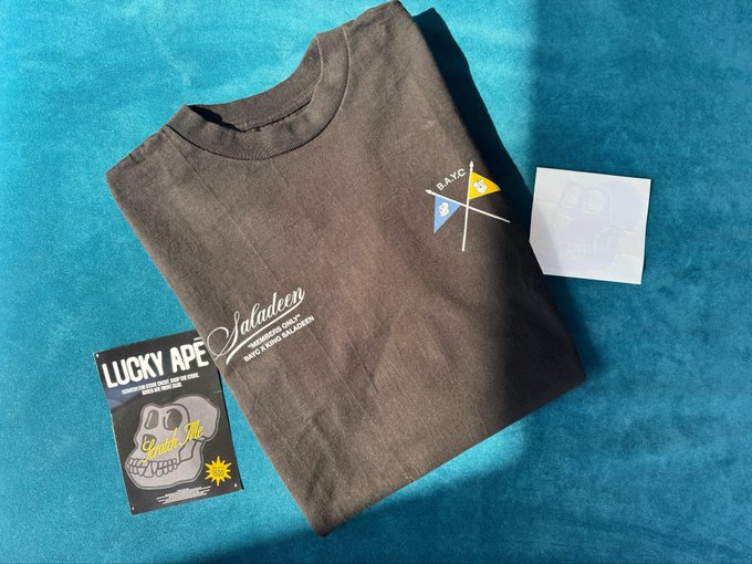 A dark grey "Saladeen" t-shirt laid out on a teal surface next to two cards, one of which reads "LUCKY APE" and features a grinning ape illustration, while the shirt bears a crossed flags graphic with "BAYC" and BAYC logos.