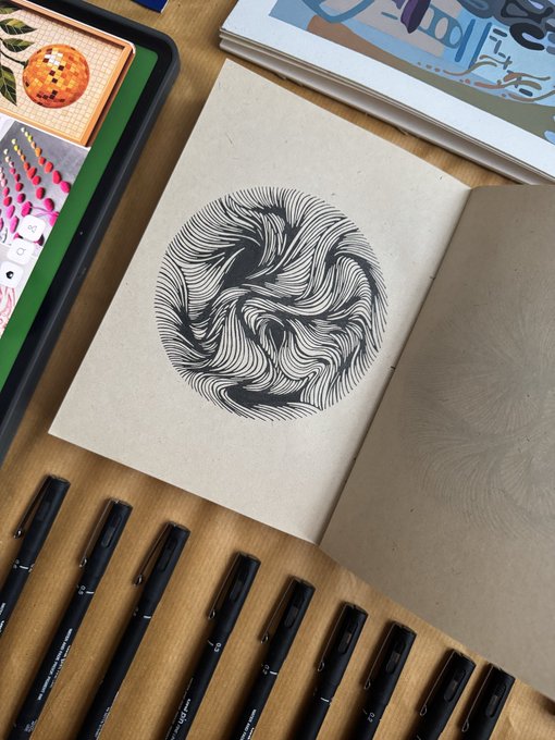 An open sketchbook displays a detailed black ink drawing of a swirling circular pattern, surrounded by black fineliner pens and colored art supplies on a wooden desk.