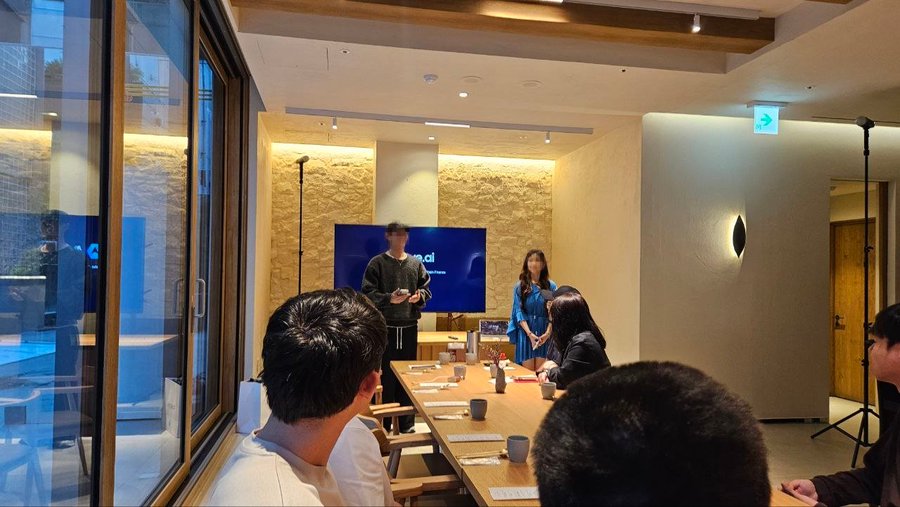 Interior shot of a presentation or meeting in a modern cafe or lounge space, with a person standing in front of a screen displaying a logo, addressing seated attendees around a wooden table.