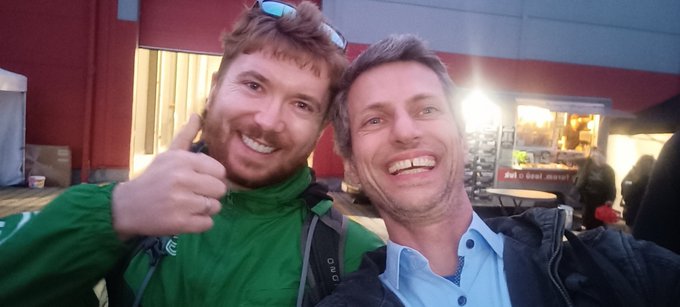 The image features two men posing for a photo, both wearing green jackets. They are standing next to each other with their arms around one another's shoulders, smiling at the camera. The background of the image shows a building, possibly an office or event venue, and there is also a truck visible in the scene. The post includes text that reads "I met Matt from @BTCPrague outside the venue of @BTCPrague." This suggests that the two men are attending an event related to Bitcoin or cryptocurrency, as indicated by the name Matt.